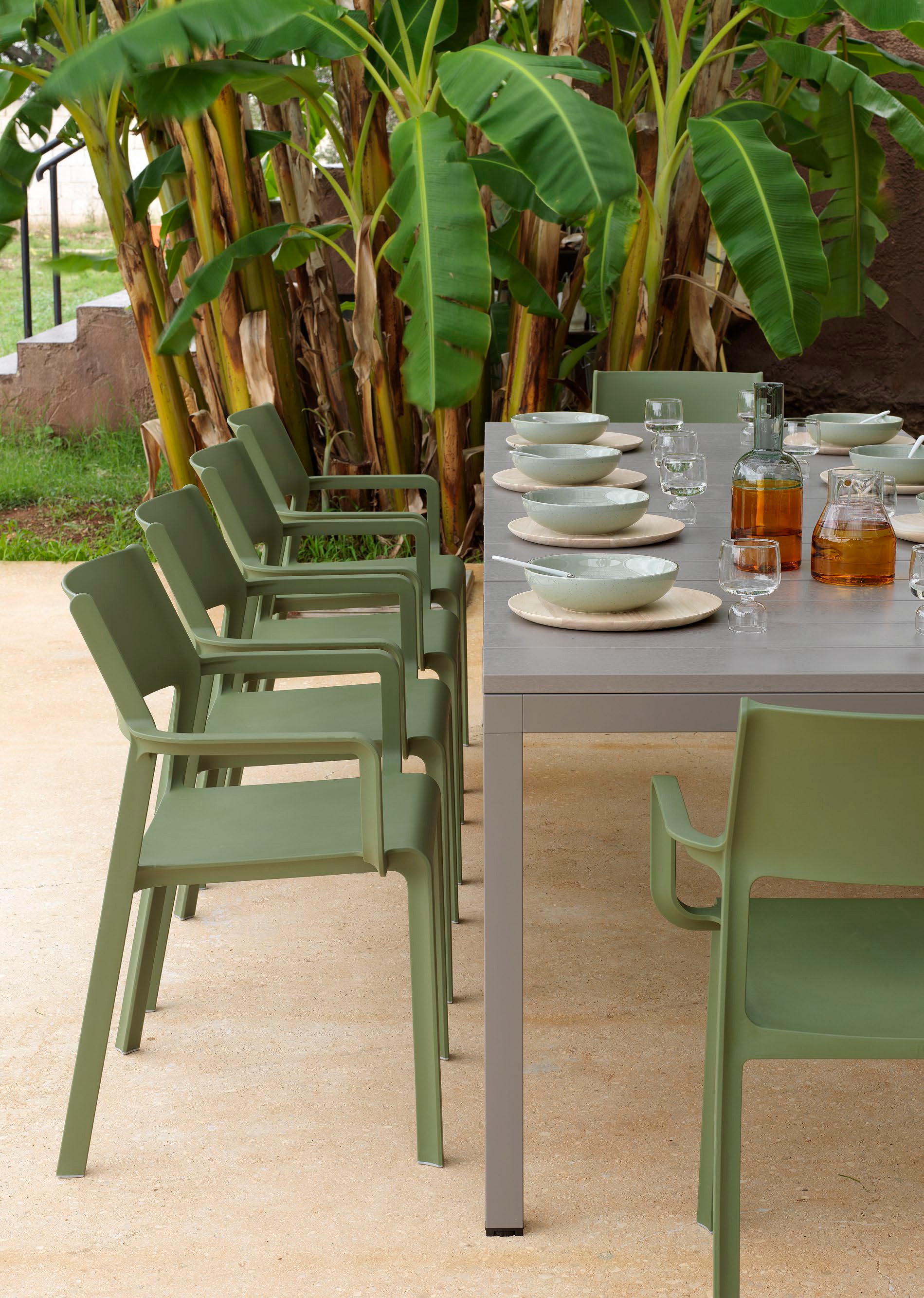 Mesa ro 210 extensible de Nardi italia color taupe sobre un piso de terraza liso, fondo de plantas y palmeras verdes. mesa decorada y servida para comer, vista frontal cortada a la mitad, con 5 sillas Trill con brazos agave.