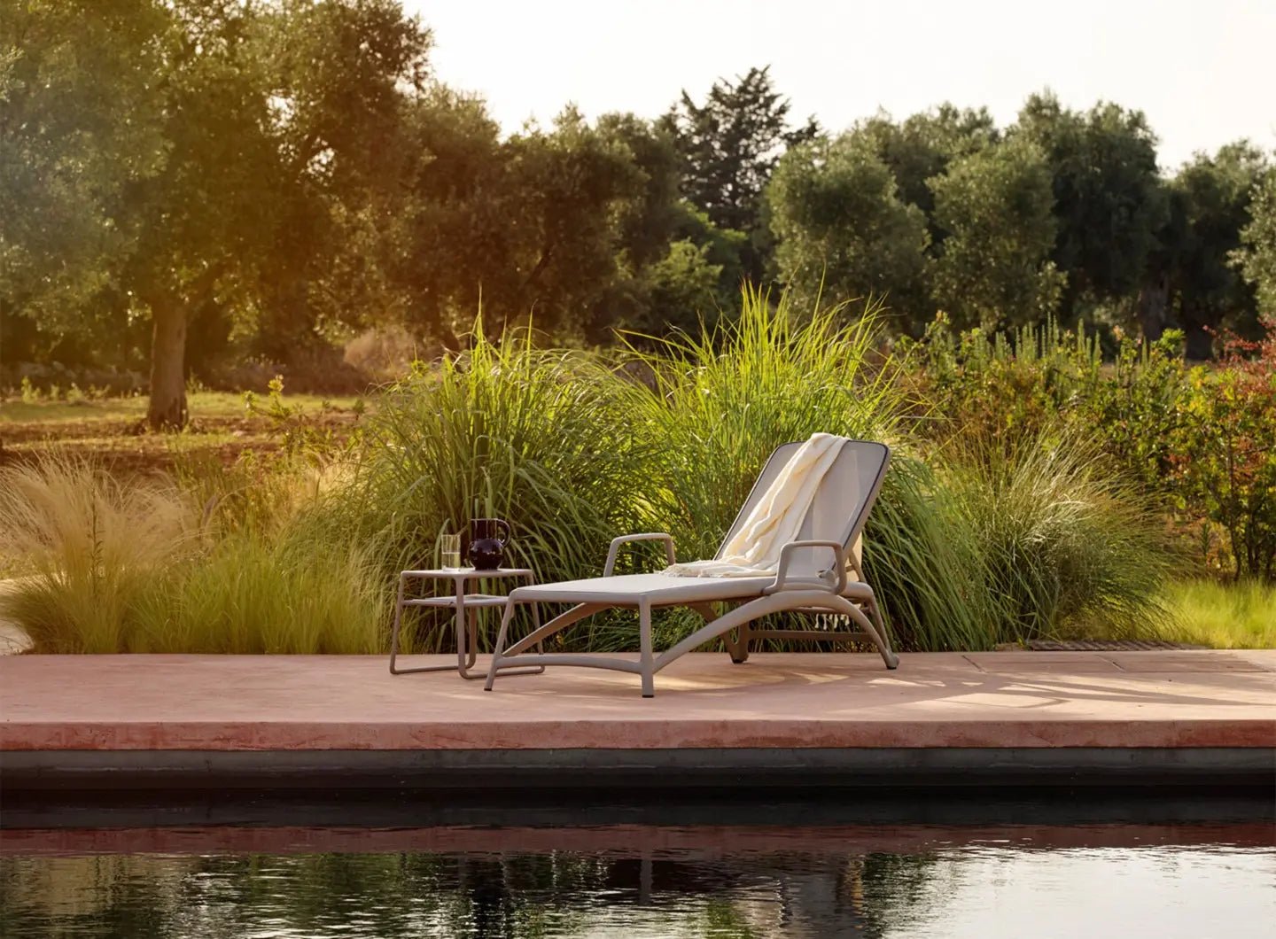 Reposera Atlántico con Apoyabrazos Nardi Italia color Tortora — ambiente junto a piscina y jardín con pastos ornamentales, al atardecer — bCasual